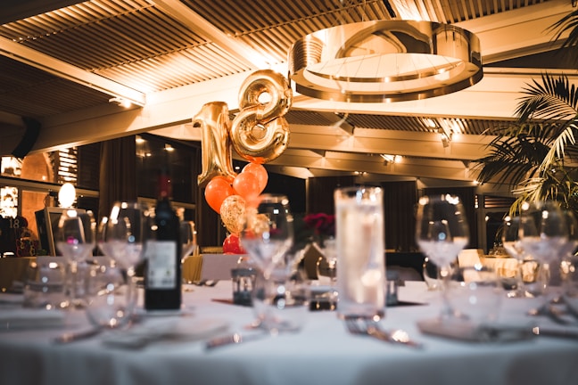 A dimly lit restaurant or event space with a large gold balloon shaped like the number 18 is surrounded by clusters of smaller orange and gold balloons. There are elegantly set tables with wine glasses, cutlery, and a bottle of wine. The ceiling features modern architectural elements and lighting, giving a warm ambiance.