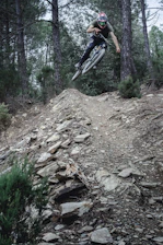 A biker speeding down a steep, rocky mountain trail surrounded by tall pine trees.