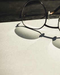 A pair of round glasses rests on a flat surface, casting a distinct shadow. The lighting creates a soft contrast between the shadows and the light-colored background, emphasizing the circular shapes of the lenses and frames.