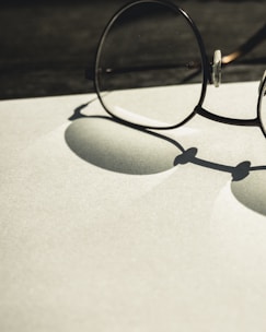 A pair of round glasses rests on a flat surface, casting a distinct shadow. The lighting creates a soft contrast between the shadows and the light-colored background, emphasizing the circular shapes of the lenses and frames.