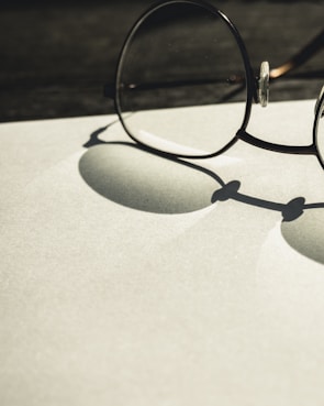 A pair of round glasses rests on a flat surface, casting a distinct shadow. The lighting creates a soft contrast between the shadows and the light-colored background, emphasizing the circular shapes of the lenses and frames.