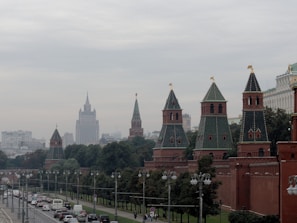 A cityscape featuring historic architecture with distinctive pointed towers and spires, likely from a significant cultural or governmental building. A busy street with traffic runs alongside a row of lush trees.
