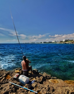 A person sits on a rock by the ocean shoreline with a fishing rod, facing the sea. Nearby, there is a cooler, a fishing tackle box, and other fishing gear. The ocean is a deep blue with waves breaking gently on the rocks. In the background, a distant skyline with buildings can be seen under a partly cloudy sky.