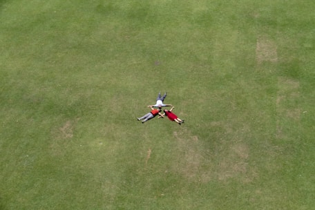 Three people are lying on a vast expanse of green grass, arranged in a circular formation with their heads meeting in the center. The setting is an open and spacious area, giving a sense of freedom and relaxation.