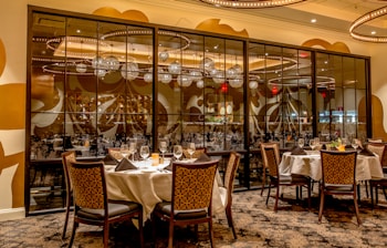 A sophisticated dining area featuring round tables dressed with white tablecloths and set with glassware, napkins, and plates. Elegant chairs surround each table. The space is illuminated by warm lighting from ornate chandeliers reflecting off large decorative mirrors adorned with artistic patterns. The atmosphere conveys a sense of luxury and refinement.