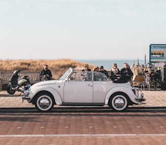 A white classic convertible car is parked on a paved area near the coast. Several people are sitting on a bench behind the car, enjoying the view of the sea. A scooter and several bicycles are also visible near a signboard.
