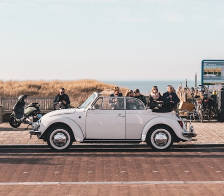 A white classic convertible car is parked on a paved area near the coast. Several people are sitting on a bench behind the car, enjoying the view of the sea. A scooter and several bicycles are also visible near a signboard.