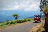 A red tuk-tuk is driving along a narrow road on a lush, green hillside. There are trees and bushes lining the road, with a scenic view of a vast valley and mountains in the background under a cloudy sky.