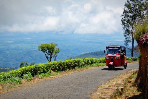 A red tuk-tuk is driving along a narrow road on a lush, green hillside. There are trees and bushes lining the road, with a scenic view of a vast valley and mountains in the background under a cloudy sky.