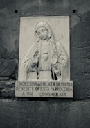 A stone relief sculpture featuring a religious figure, likely the Virgin Mary, with a serene expression. The figure holds hands in a gesture of blessing and has a heart motif on the chest. Below the sculpture, there is an Italian inscription carved into the stone. The surrounding wall has a rough, textured surface.