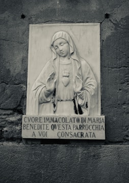 A stone relief sculpture featuring a religious figure, likely the Virgin Mary, with a serene expression. The figure holds hands in a gesture of blessing and has a heart motif on the chest. Below the sculpture, there is an Italian inscription carved into the stone. The surrounding wall has a rough, textured surface.