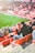 A group of people is sitting in bright red stadium seats, watching a live sports event. The stadium is well-lit with natural sunlight, and the grass on the field is vivid green. Most spectators are dressed in casual, warm clothing, reflecting cooler weather. The crowd is engaged, with many spectators clapping and attentive to the event.