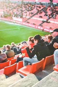 A group of people is sitting in bright red stadium seats, watching a live sports event. The stadium is well-lit with natural sunlight, and the grass on the field is vivid green. Most spectators are dressed in casual, warm clothing, reflecting cooler weather. The crowd is engaged, with many spectators clapping and attentive to the event.