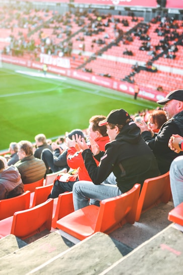 A group of people is sitting in bright red stadium seats, watching a live sports event. The stadium is well-lit with natural sunlight, and the grass on the field is vivid green. Most spectators are dressed in casual, warm clothing, reflecting cooler weather. The crowd is engaged, with many spectators clapping and attentive to the event.