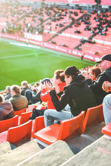 A group of people is sitting in bright red stadium seats, watching a live sports event. The stadium is well-lit with natural sunlight, and the grass on the field is vivid green. Most spectators are dressed in casual, warm clothing, reflecting cooler weather. The crowd is engaged, with many spectators clapping and attentive to the event.