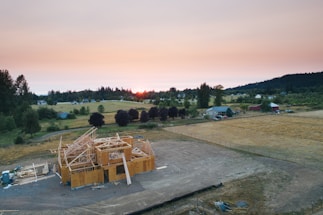 Aerial view of a partially constructed house surrounded by open fields and distant trees at sunset. The structure is framed with wooden beams, and there are scattered construction materials nearby. The landscape features a mix of green and yellow vegetation with a few residential houses and farm buildings in the distance.