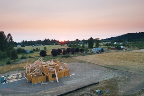 Aerial view of a partially constructed house surrounded by open fields and distant trees at sunset. The structure is framed with wooden beams, and there are scattered construction materials nearby. The landscape features a mix of green and yellow vegetation with a few residential houses and farm buildings in the distance.