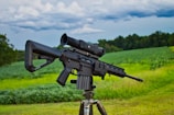 A black rifle equipped with a scope is mounted on a tripod, positioned in a lush green field with trees in the background. The sky above is cloudy, suggesting overcast weather.