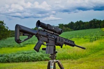 A black rifle equipped with a scope is mounted on a tripod, positioned in a lush green field with trees in the background. The sky above is cloudy, suggesting overcast weather.