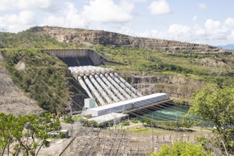 A large hydroelectric power plant located in a hilly landscape, featuring multiple penstocks leading into a powerhouse. The structure spans a river with visible power lines and surrounding greenery.