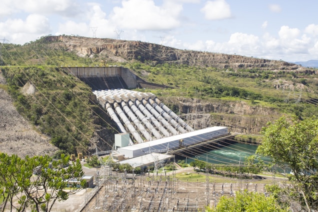 A large hydroelectric power plant located in a hilly landscape, featuring multiple penstocks leading into a powerhouse. The structure spans a river with visible power lines and surrounding greenery.