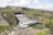A large hydroelectric power plant located in a hilly landscape, featuring multiple penstocks leading into a powerhouse. The structure spans a river with visible power lines and surrounding greenery.