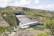 A large hydroelectric power plant located in a hilly landscape, featuring multiple penstocks leading into a powerhouse. The structure spans a river with visible power lines and surrounding greenery.