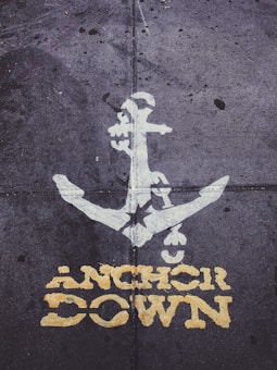 A white anchor symbol is painted on a textured, gray concrete surface. Below the anchor, the words 'ANCHOR DOWN' are stenciled in bold, yellow lettering. The overall appearance suggests a rugged, urban setting.