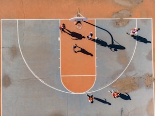 A top-down view of a basketball court with several players engaged in a game. The court features a distinctive orange and blue color scheme, and the shadows cast by the players suggest a sunny day. The basketball hoop, lines of the court, and multiple players in motion are clearly visible.