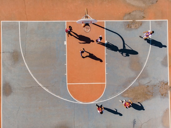 A top-down view of a basketball court with several players engaged in a game. The court features a distinctive orange and blue color scheme, and the shadows cast by the players suggest a sunny day. The basketball hoop, lines of the court, and multiple players in motion are clearly visible.