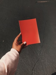 A person's hand holding a bright red piece of paper over a textured gray surface, such as a paved street. The hand is positioned centrally, and the paper contrasts starkly against the darker background.