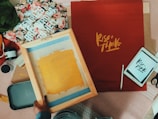 A creative workspace with various artistic tools and materials spread out. A screen printing frame with a yellow base layer occupies the center, while a red fabric with the text 'Rise + Thrive' printed in bold yellow lettering is placed nearby. Floral fabric and a tablet displaying similar text are also visible. Additional items include paint containers, brushes, and fabric swatches.