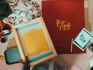 A creative workspace with various artistic tools and materials spread out. A screen printing frame with a yellow base layer occupies the center, while a red fabric with the text 'Rise + Thrive' printed in bold yellow lettering is placed nearby. Floral fabric and a tablet displaying similar text are also visible. Additional items include paint containers, brushes, and fabric swatches.