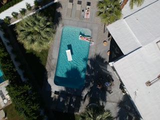 Aerial view of a swimming pool surrounded by palm trees and a modern building with a flat roof. Two people are sunbathing on loungers beside the pool, and the bright sunlight creates distinct shadows. The area is landscaped with greenery and gravel paths.