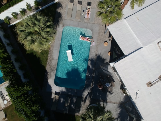 Aerial view of a swimming pool surrounded by palm trees and a modern building with a flat roof. Two people are sunbathing on loungers beside the pool, and the bright sunlight creates distinct shadows. The area is landscaped with greenery and gravel paths.