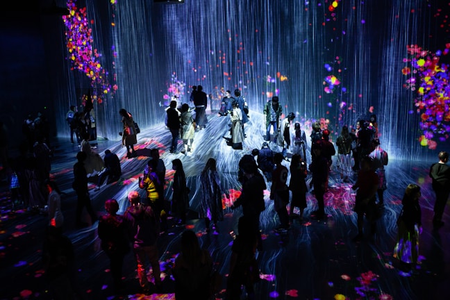 A crowd of people stands in a dark room illuminated by a vibrant digital projection. The projection features cascading lines resembling a waterfall, interspersed with colorful patches resembling flowers or fireworks. The light reflects off the floor, creating an immersive and interactive environment.