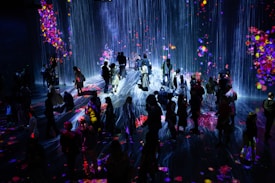 A crowd of people stands in a dark room illuminated by a vibrant digital projection. The projection features cascading lines resembling a waterfall, interspersed with colorful patches resembling flowers or fireworks. The light reflects off the floor, creating an immersive and interactive environment.