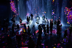 A crowd of people stands in a dark room illuminated by a vibrant digital projection. The projection features cascading lines resembling a waterfall, interspersed with colorful patches resembling flowers or fireworks. The light reflects off the floor, creating an immersive and interactive environment.