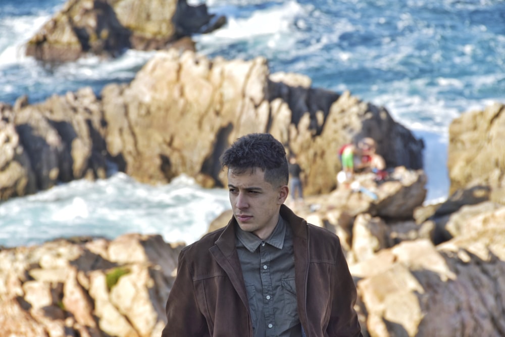 man wearing brown leather jacket standing on rock formation near the sea during daytime