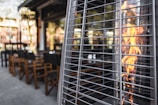 An outdoor patio setting with wooden chairs and tables near a tall outdoor heater with flames visible through its metal grid. The patio area is surrounded by rows of buildings and greenery is subtly reflected in the background.