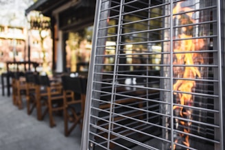 An outdoor patio setting with wooden chairs and tables near a tall outdoor heater with flames visible through its metal grid. The patio area is surrounded by rows of buildings and greenery is subtly reflected in the background.