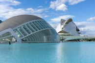 Modern architectural structures are seen next to a large body of water under a blue sky with scattered clouds. The buildings have a futuristic design with curved roofs and glass panels, and a person is standing next to the water, possibly for maintenance.