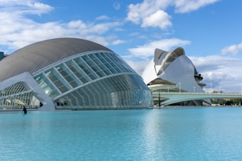 Modern architectural structures are seen next to a large body of water under a blue sky with scattered clouds. The buildings have a futuristic design with curved roofs and glass panels, and a person is standing next to the water, possibly for maintenance.