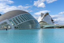 Modern architectural structures are seen next to a large body of water under a blue sky with scattered clouds. The buildings have a futuristic design with curved roofs and glass panels, and a person is standing next to the water, possibly for maintenance.