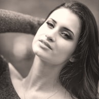 A black and white portrait of a woman with long hair. She gazes softly at the camera with her head slightly tilted. The expression on her face is calm and serene, and she has one hand resting behind her head. Her features are highlighted by the use of shadow and light, creating a smooth and elegant look.
