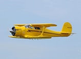 A vintage yellow aircraft is flying against a clear blue sky. It features a distinctive radial engine and a streamlined fuselage with black and red accents. The registration number N9405H is visible on the tail.