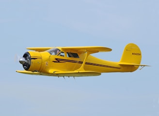 A vintage yellow aircraft is flying against a clear blue sky. It features a distinctive radial engine and a streamlined fuselage with black and red accents. The registration number N9405H is visible on the tail.