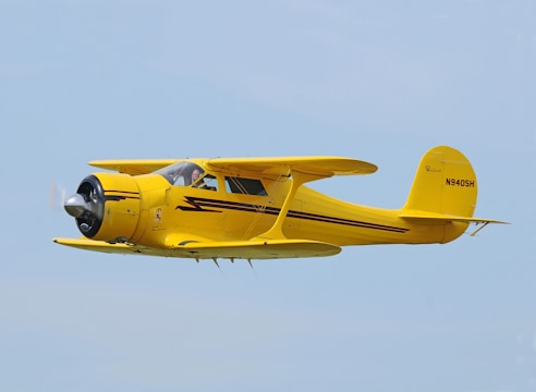 A vintage yellow aircraft is flying against a clear blue sky. It features a distinctive radial engine and a streamlined fuselage with black and red accents. The registration number N9405H is visible on the tail.