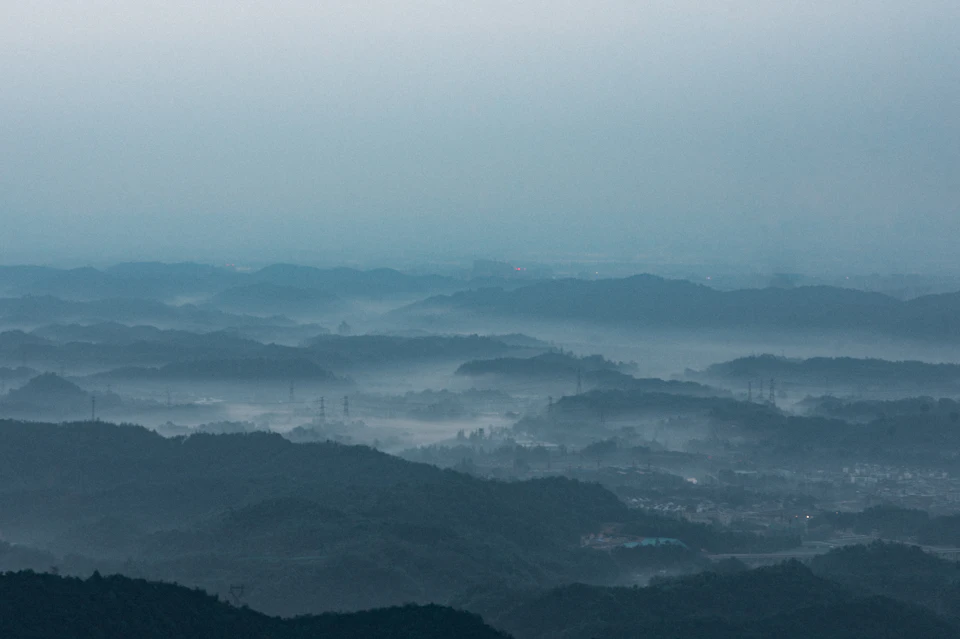 The image depicts a breathtaking view from a high vantage point, overlooking a vast landscape of rolling hills and mountains blanketed in fog. The sky above is a light blue canvas, dotted with wispy clouds that add depth to the scene. In the distance, several buildings can be seen nestled among the natural beauty of the landscape.