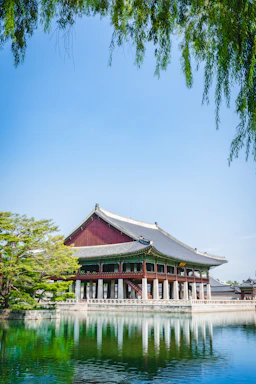 A traditional Korean building with a curved roof stands on the edge of a reflective pond, surrounded by lush greenery. The clear blue sky provides a serene backdrop, while willow branches hang gracefully at the top.